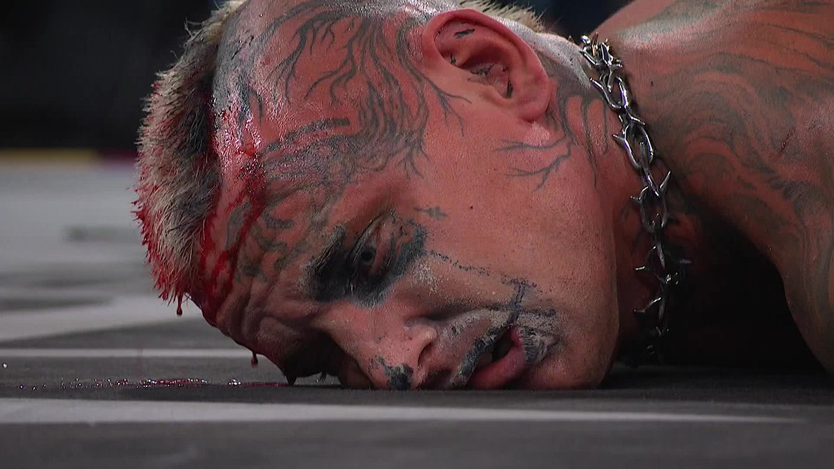 A close-up shows professional wrestler Darby Allin lying face-down on the mat, his face covered in blood and black face paint smeared around his eyes and mouth, with a chain draped around his neck—visually capturing the brutal beating described in the caption as Kyle Fletcher bashes in his skull.