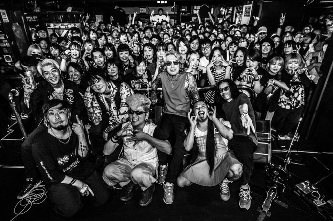 A black and white photograph showing a large group of fans posing with musicians at what appears to be a concert venue, with a 'TOILET' sign visible in the background.