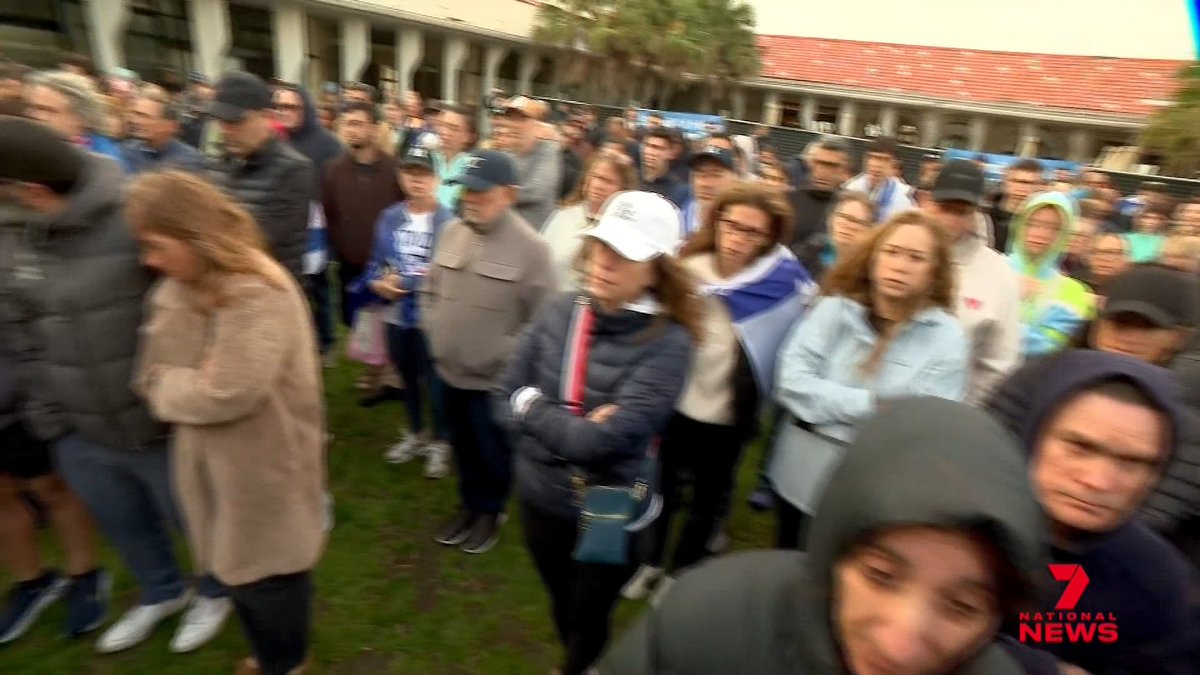 A Channel 7 National News screenshot showing a crowd of people gathered outdoors in cold weather near a stadium with red seating.