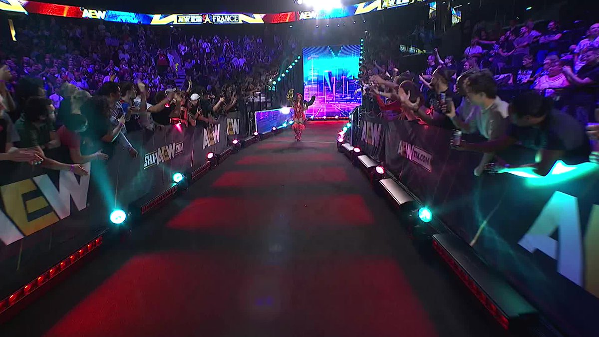 A wrestler makes her entrance down a red-carpeted ramp lined with cheering fans, while illuminated AEW signage and a cityscape backdrop set the stage for the #AEWGrandSlam event in France.