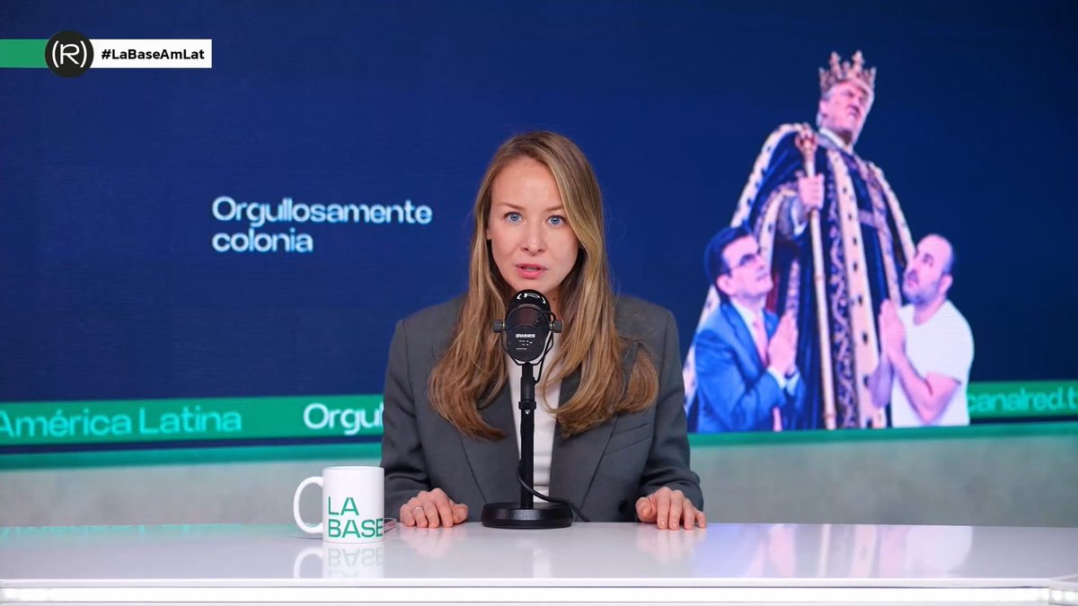 A female news anchor in a gray blazer at a desk with a 'LA BASE' mug and microphone, with the broadcast graphic showing '#LaBaseAmLat' and text reading 'Orgullosamente colonia' alongside an image of a crowned figure being carried.