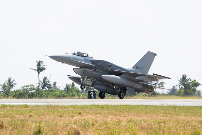 A gray F-16 fighter jet, marked with the number '5727', takes off from a runway with palm trees visible in the background.