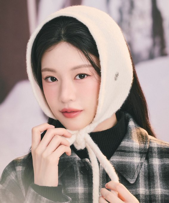 A portrait of a young woman with long dark hair wearing a white fluffy hood and a plaid jacket over a black turtleneck.