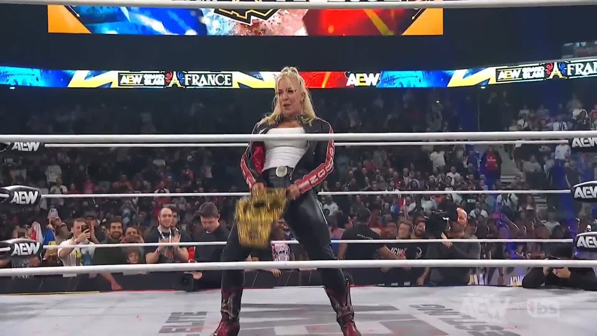 A female wrestler with blonde hair in a ponytail stands in an AEW wrestling ring at an AEW x NJPW Forbidden Door event in France, holding a championship belt.