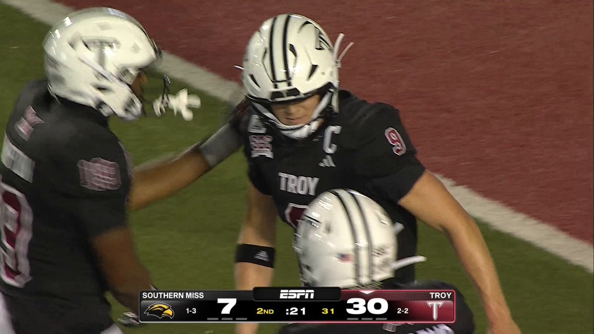 Two Troy Trojans players celebrate on the field during their dominant 30-7 lead over Southern Miss with just 21 seconds left in the 2nd quarter, with one player wearing the captain's "C" designation and number 9 visible. The scoreboard at the bottom of the ESPN broadcast shows Troy (2-2) controlling the game against Southern Miss (1-3), reflecting the team's earlier success executing a "Spider 2 Y Banana" play on a crucial 4th and 1 while up 24-7.