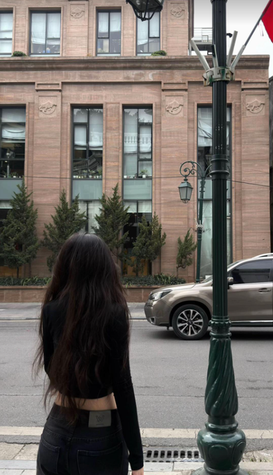 A woman with long dark hair, wearing a black outfit, faces a city street where an SUV drives past a brick building with large windows and a decorative green lamppost in the foreground.