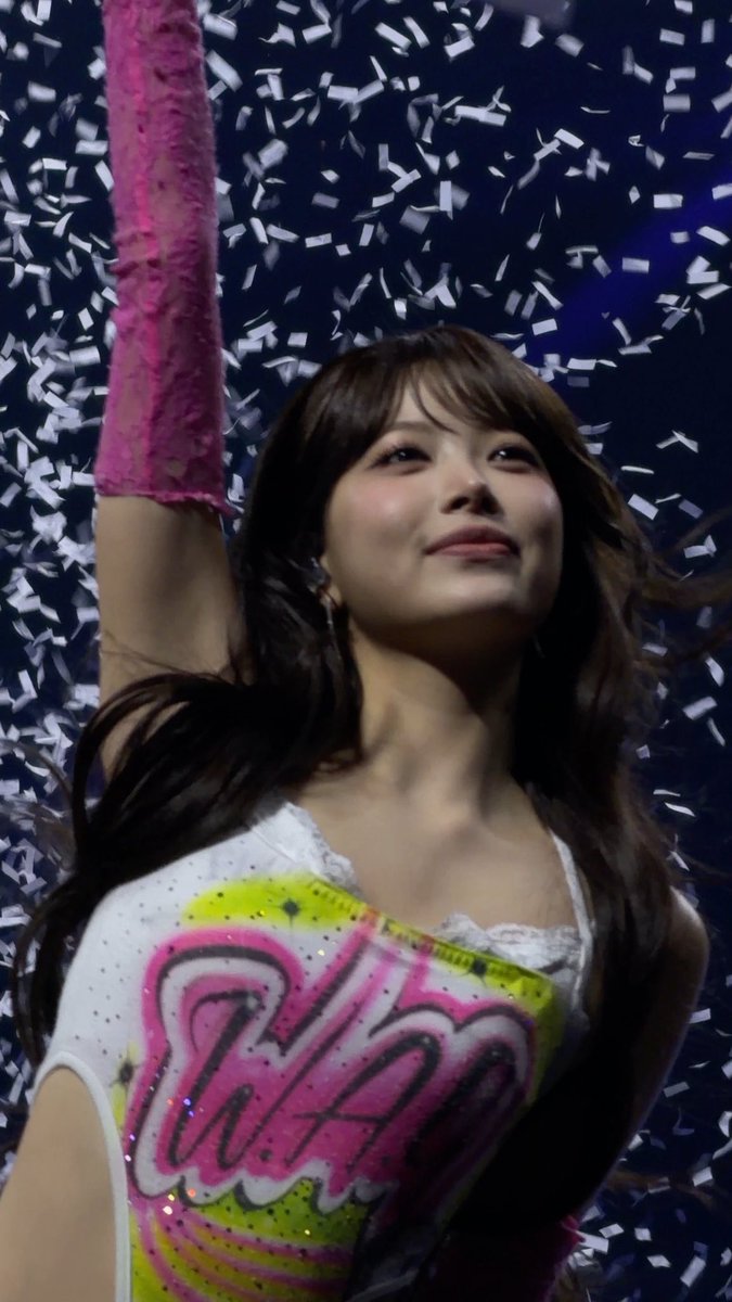A woman in a colorful outfit with "Hi" written on it smiles and raises her arm as confetti falls around her.