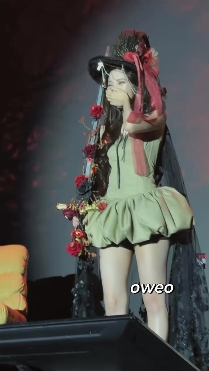 A performer in a green dress and dark floral headpiece stands on stage covering her mouth with her hand, with the text 'oweo' visible in the corner.
