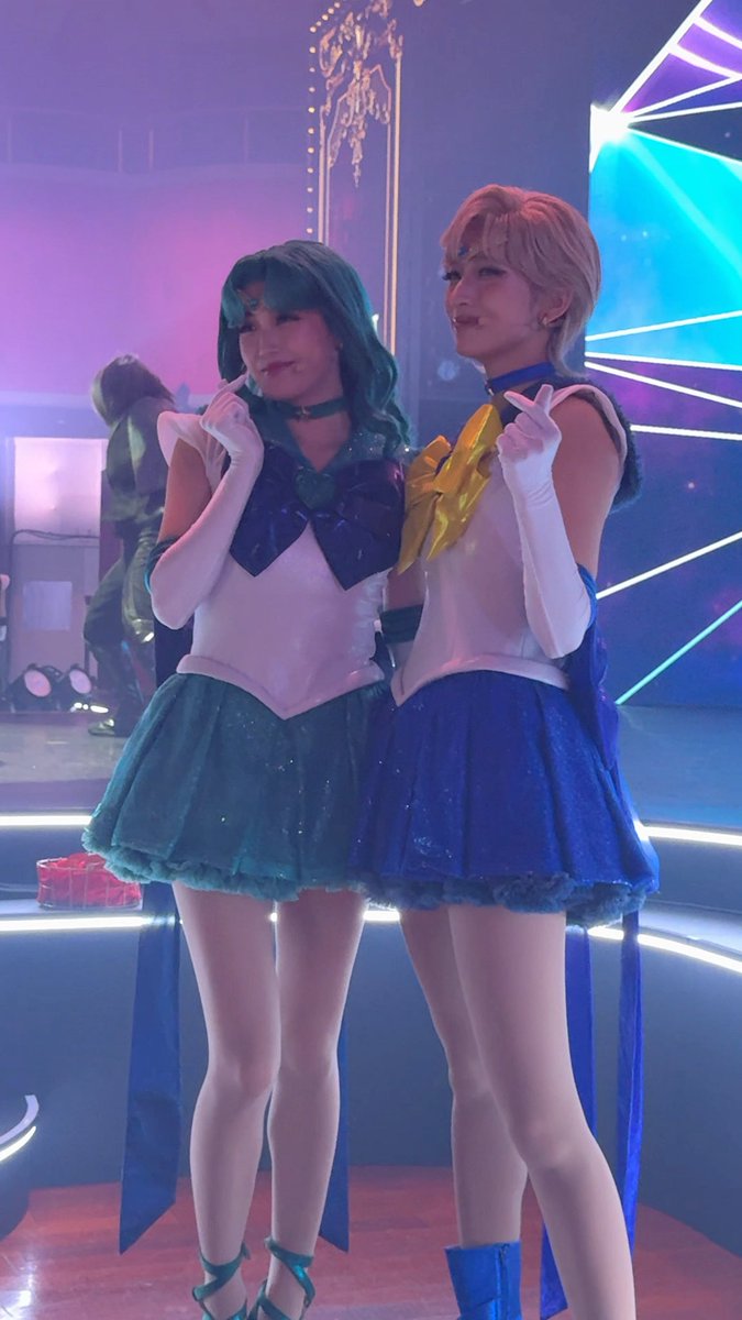 Two individuals in Sailor Moon cosplay, dressed in blue and white outfits with skirts and gloves, pose together on a stage.