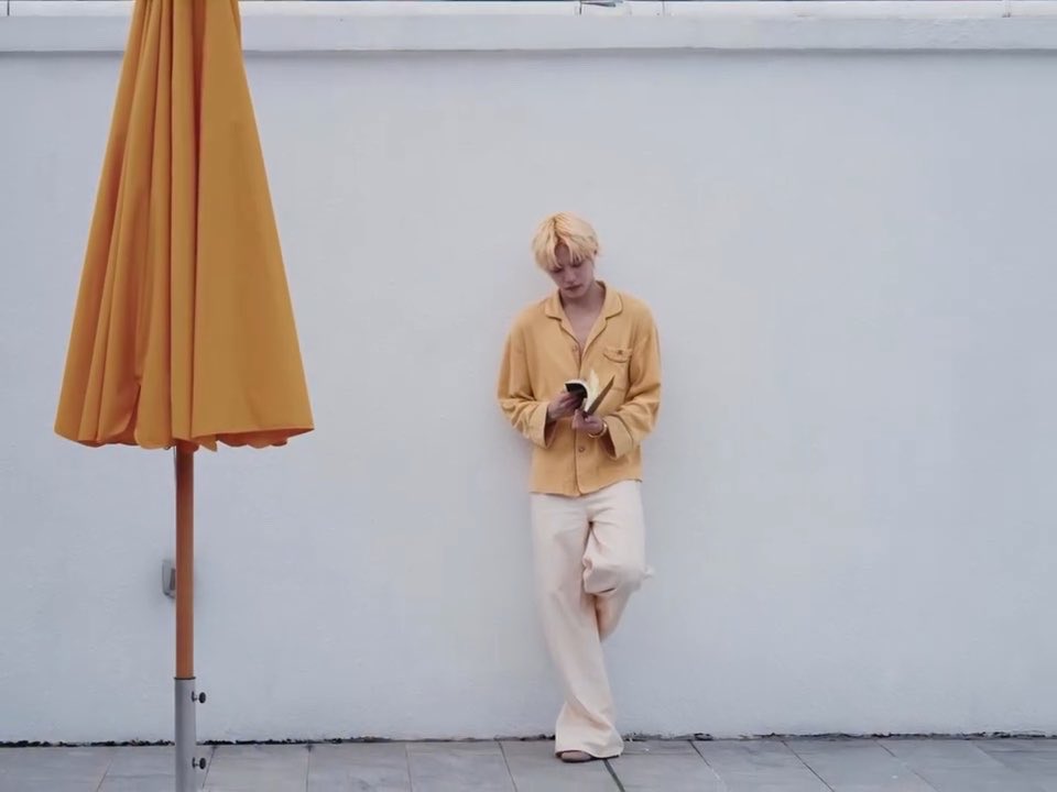 A person with blonde hair wearing a yellow shirt and white pants leans against a white wall reading a book, next to a closed yellow umbrella.
