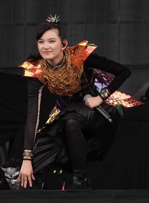 A performer in an elaborate stage costume with gold and iridescent orange shoulder pieces crouches on stage holding a microphone.