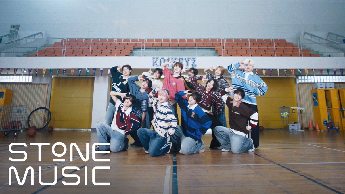 A group of thirteen young men posing playfully in a gymnasium with wooden floors and orange bleachers, with the text "STONE MUSIC" in the lower left corner.