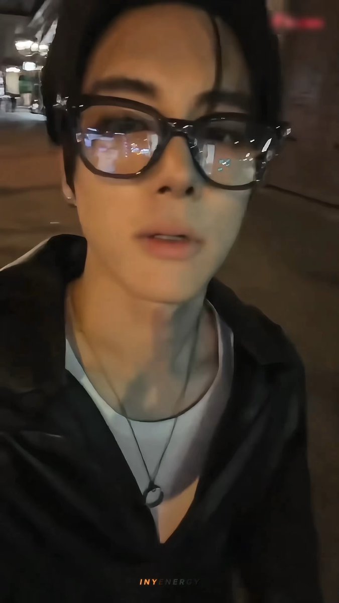 A selfie of a person wearing large tortoiseshell glasses, a black jacket over a white shirt, with a necklace, taken in a dimly lit area with the watermark 'INYENERGY'.