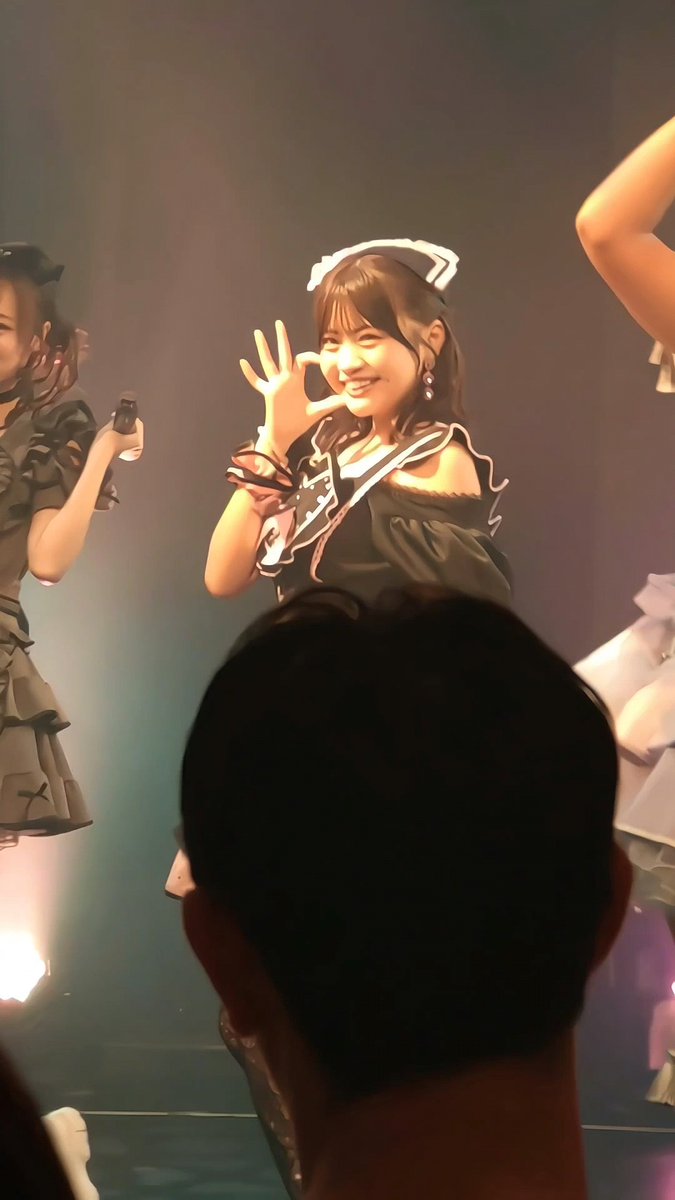 A young woman on stage at a performance making an 'OK' hand gesture, wearing a black and white outfit with a headband, with other performers visible nearby.