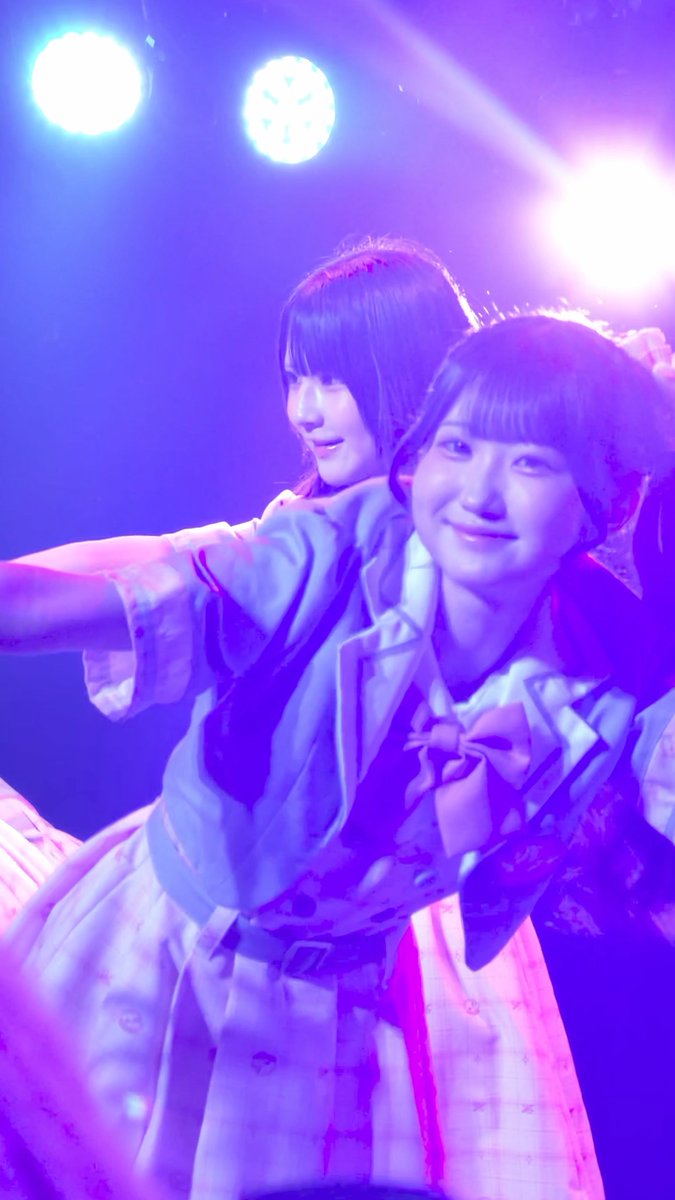 Two young women in coordinated outfits perform on stage under purple and white spotlights, smiling and posing for the audience.