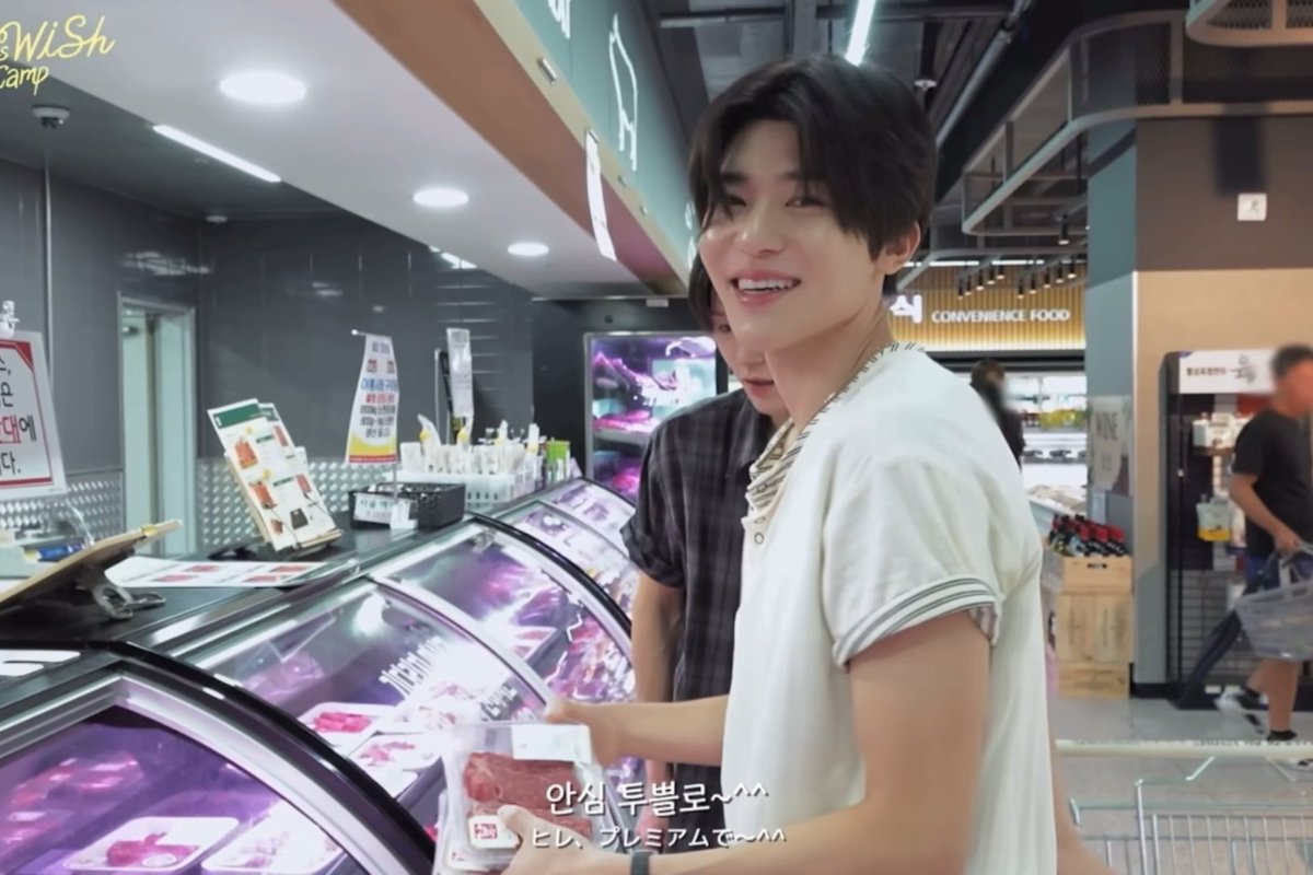 A young man in a white shirt smiles while shopping at a Korean convenience store/mart with meat displays, holding a package labeled "한우" (Hanwoo beef), with subtitles reading "안심 투블로~^^" and "티. R. 프리미엄으로~^^".