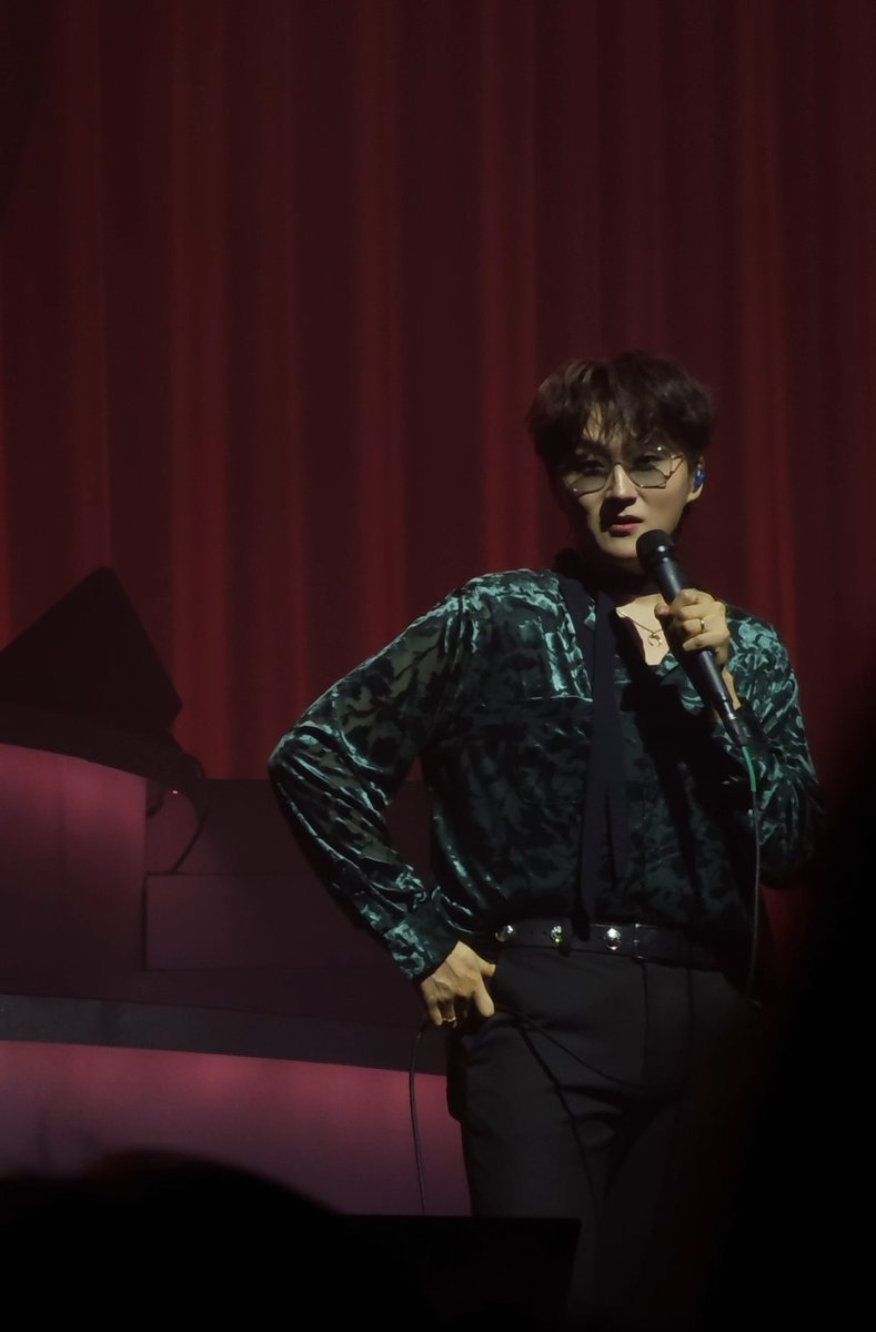 A performer wearing glasses and a dark green patterned velvet shirt stands on stage holding a microphone against a red curtain backdrop.