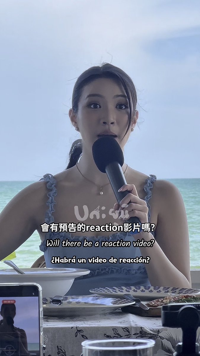 Screenshot of a video where a woman in a blue floral top speaks into a microphone on a beach, with subtitles in Chinese, English ('Will there be a reaction video?'), and Spanish ('¿Habrá un video de reacción?').
