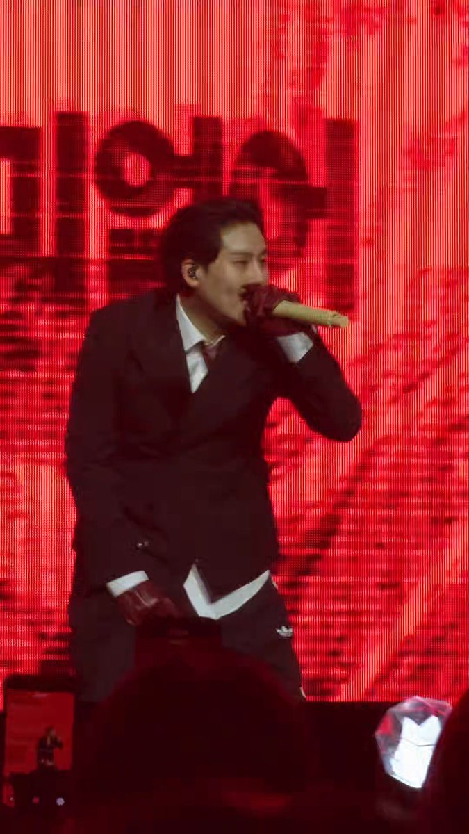 A male performer in a black suit and red gloves sings into a gold microphone on stage against a large red LED backdrop.