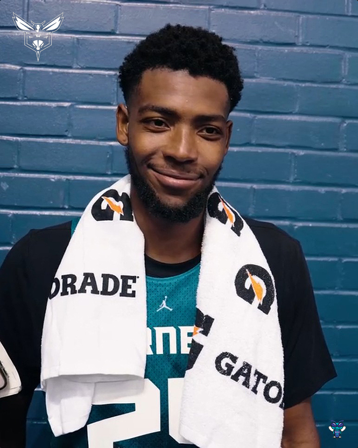 A young man wearing a teal basketball jersey with a Gatorade towel draped over his shoulders, smiling at the camera against a blue brick wall.