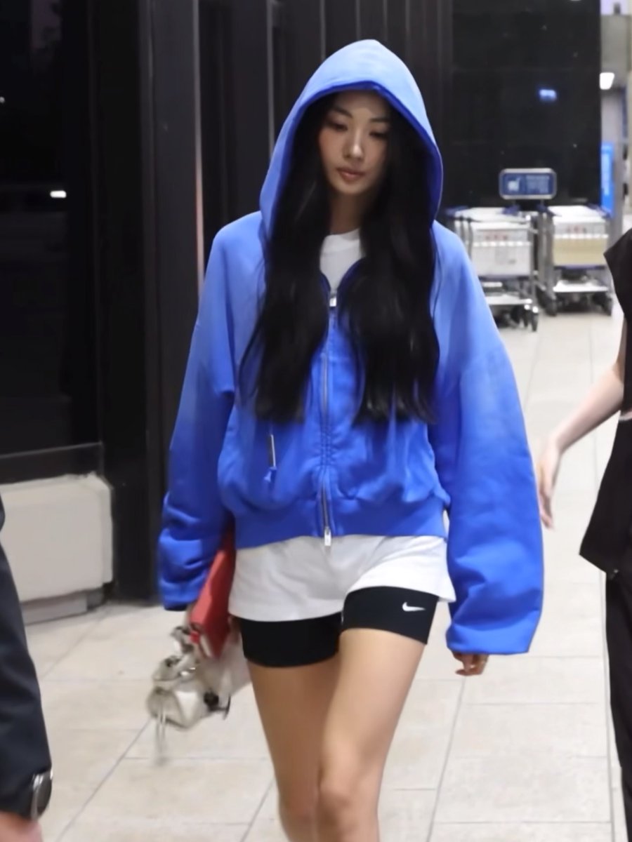 A woman with long dark hair walks through what appears to be an airport or transit area, wearing an oversized bright blue hoodie, white top, and black Nike shorts.