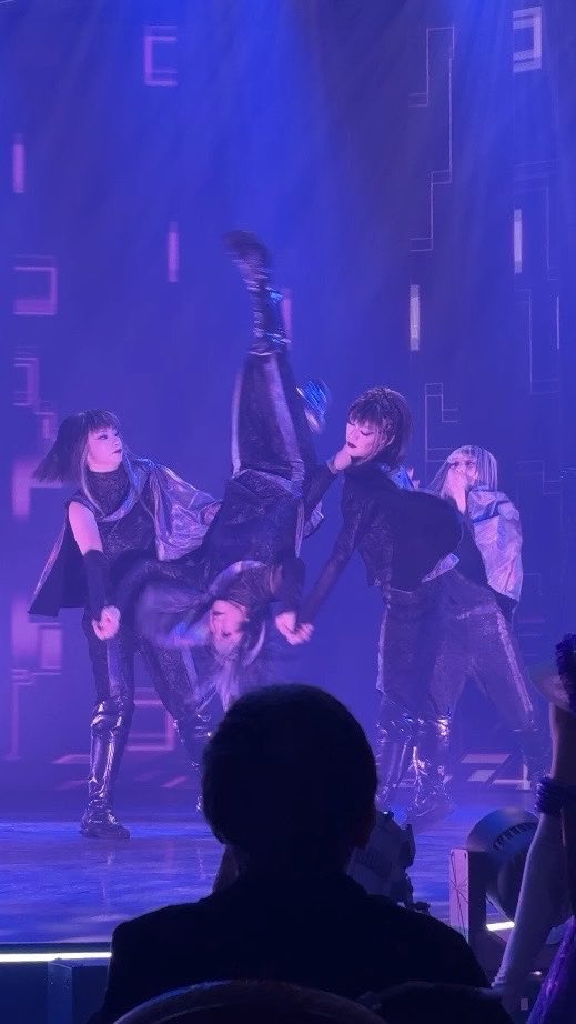 A group of performers on a stage illuminated by purple lighting, with one dancer doing a handstand supported by others.