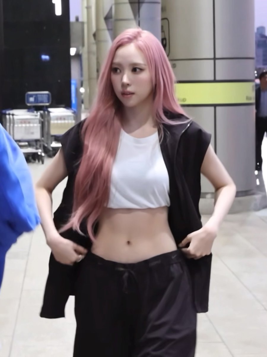 A woman with long pink hair poses in what appears to be an airport terminal, wearing a white crop top and an open black sleeveless vest, adjusting her waistband.
