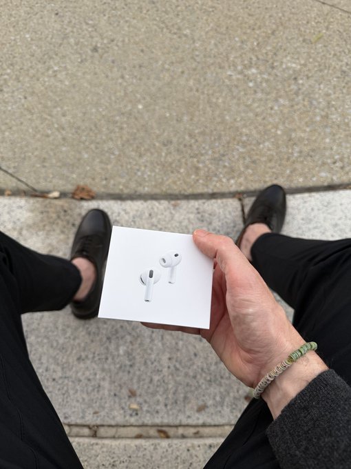 A person wearing black shoes and pants holds a white AirPods box in their hand while standing on a concrete surface, wearing a green hair tie on their wrist.