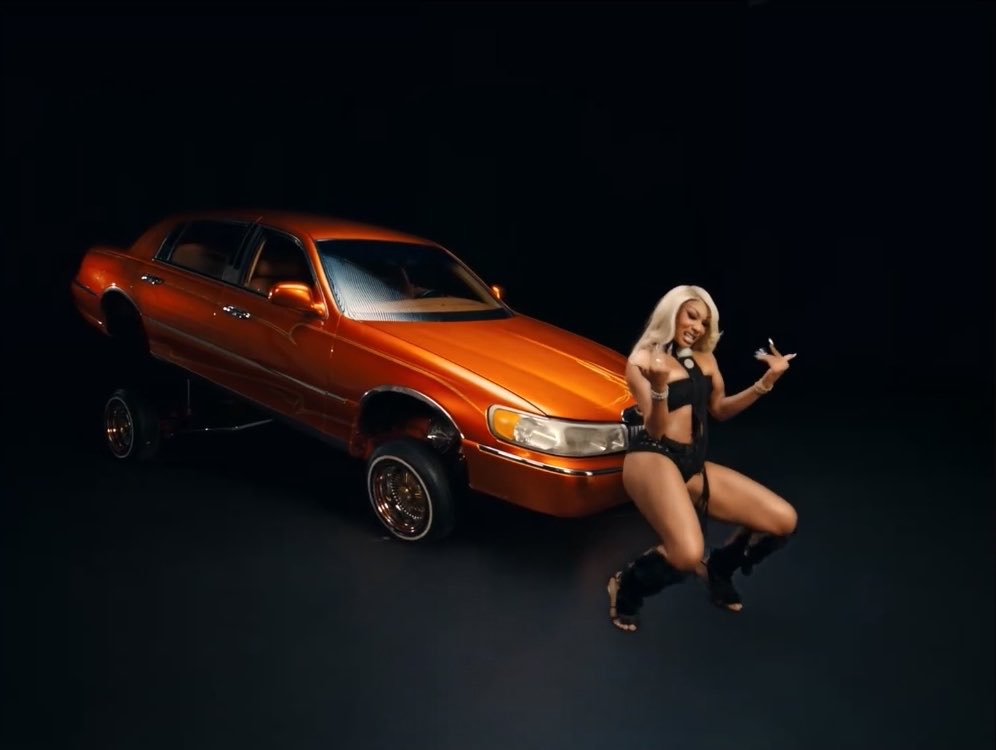 A woman with blonde hair poses in a black outfit next to a customized orange lowrider car on hydraulics against a dark background.