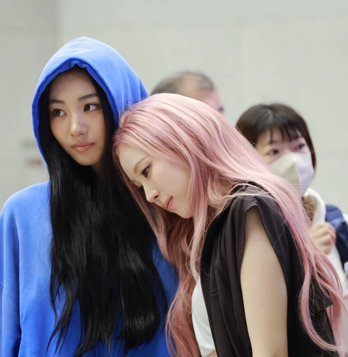 Two women standing close together; the woman on the left wears a blue hoodie and has long dark hair, while the woman on the right leans against her, sporting long pink hair and a black vest.