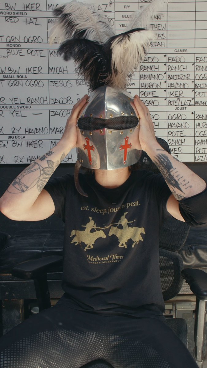 A person wearing a knight's helmet with feathers, sitting in front of a whiteboard with tournament schedules at Medieval Times, wearing a black t-shirt that reads 'eat. sleep. joust. repeat.'