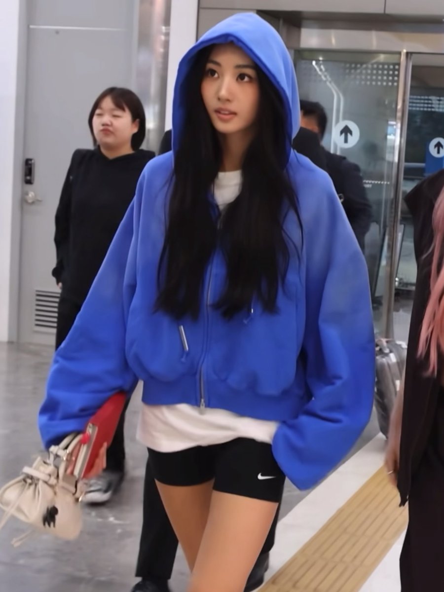 A young woman with long black hair wearing an oversized blue zip-up hoodie and black Nike shorts walks through what appears to be an airport terminal.