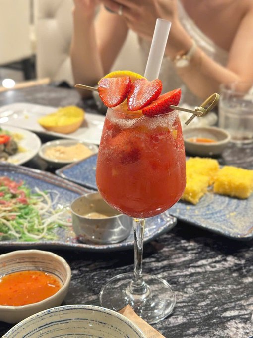 A pink strawberry cocktail garnished with fresh strawberries and a lime slice sits on a table surrounded by various small dishes of food.