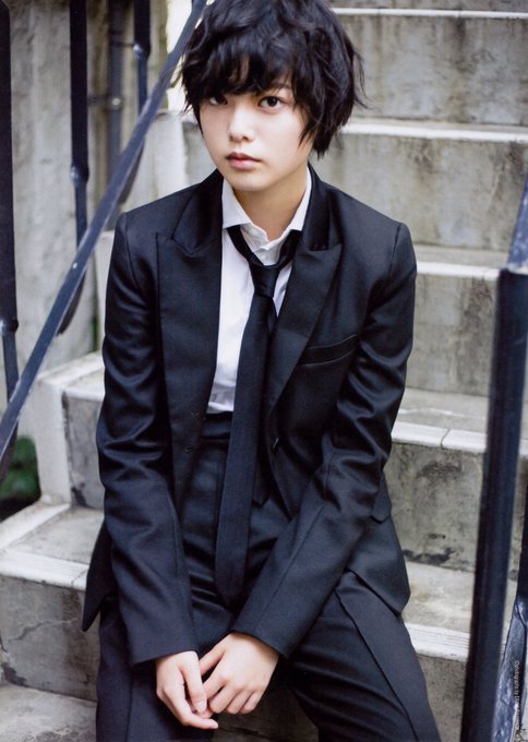 A young man with messy black hair wearing a black suit, white shirt, and black tie, sitting on stone steps.