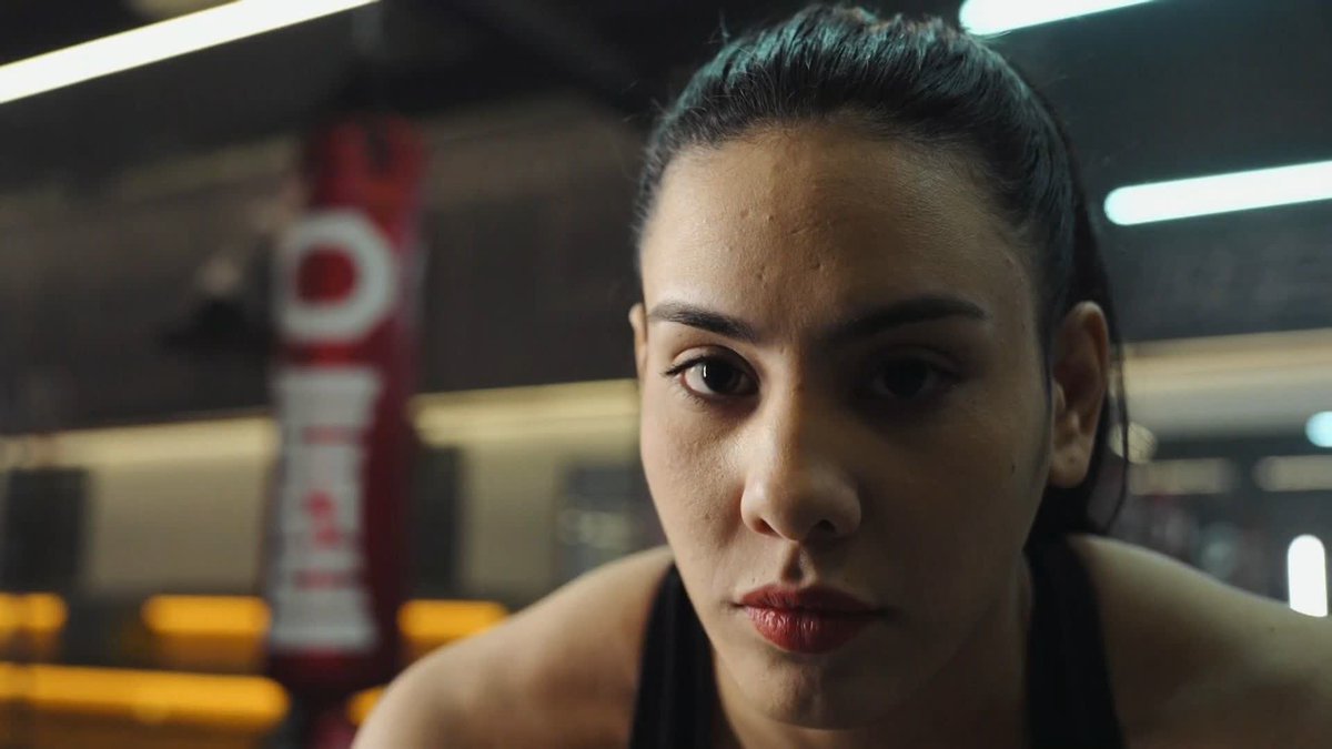 Camila Reynoso, an Argentine fighter, is shown in a determined close-up inside what appears to be a boxing gym, with a red "DOLLAR" punching bag visible behind her. Her intense gaze and athletic setting reflect her ambition to chase her dream and prove that anything can be achieved in the cage.