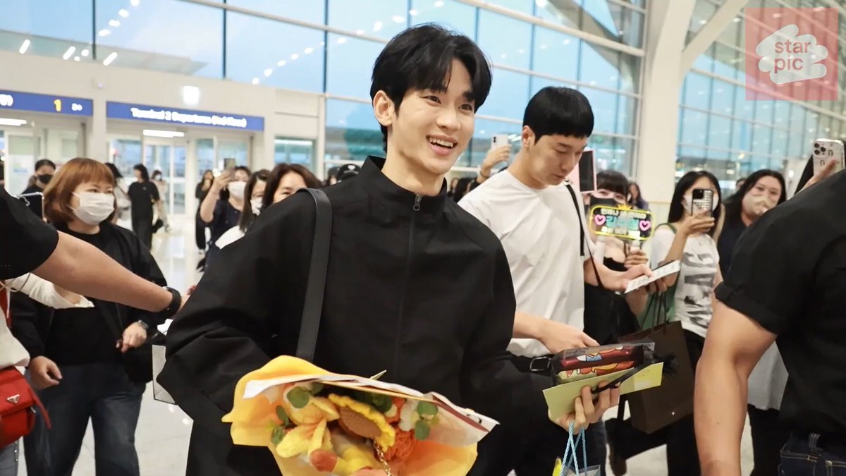 A young man in a black jacket smiles while holding items at what appears to be an airport terminal, surrounded by fans taking photos; the image bears a "starpic" watermark.