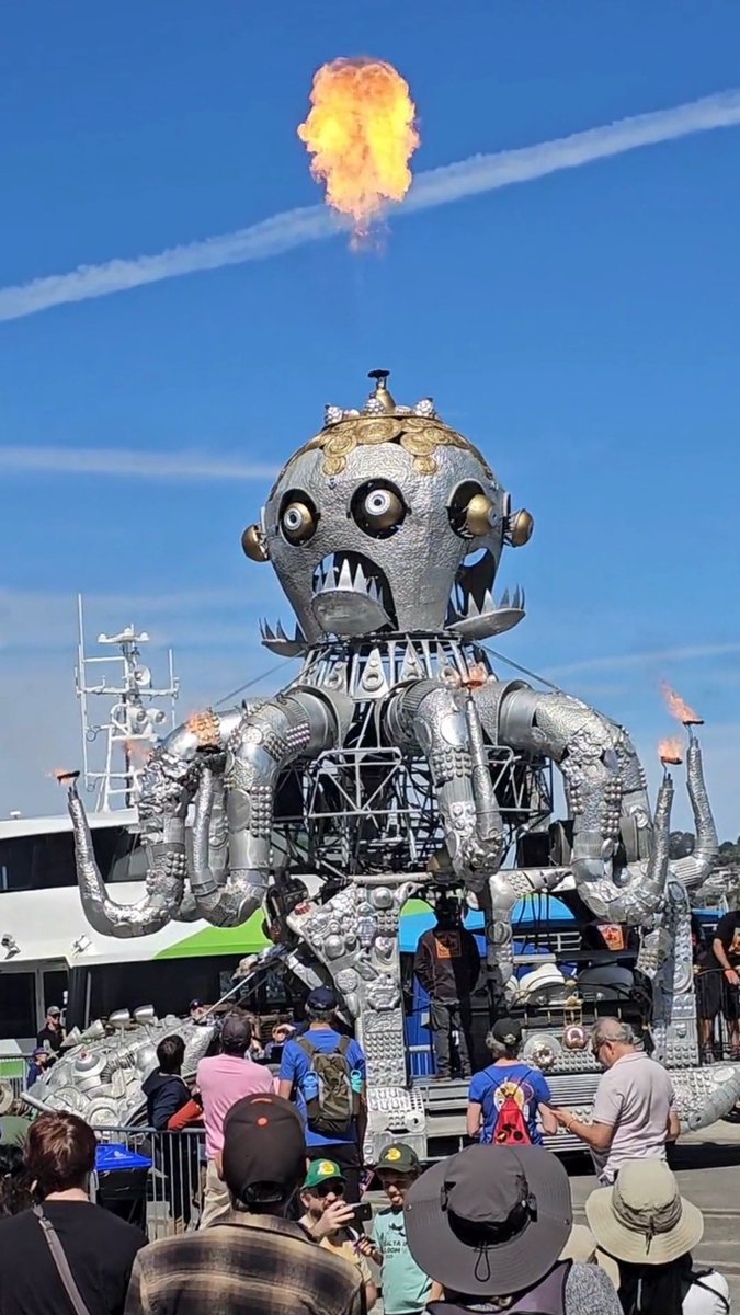 A large mechanical octopus sculpture shoots a plume of fire into the blue sky at an outdoor festival, with spectators watching in the foreground.
