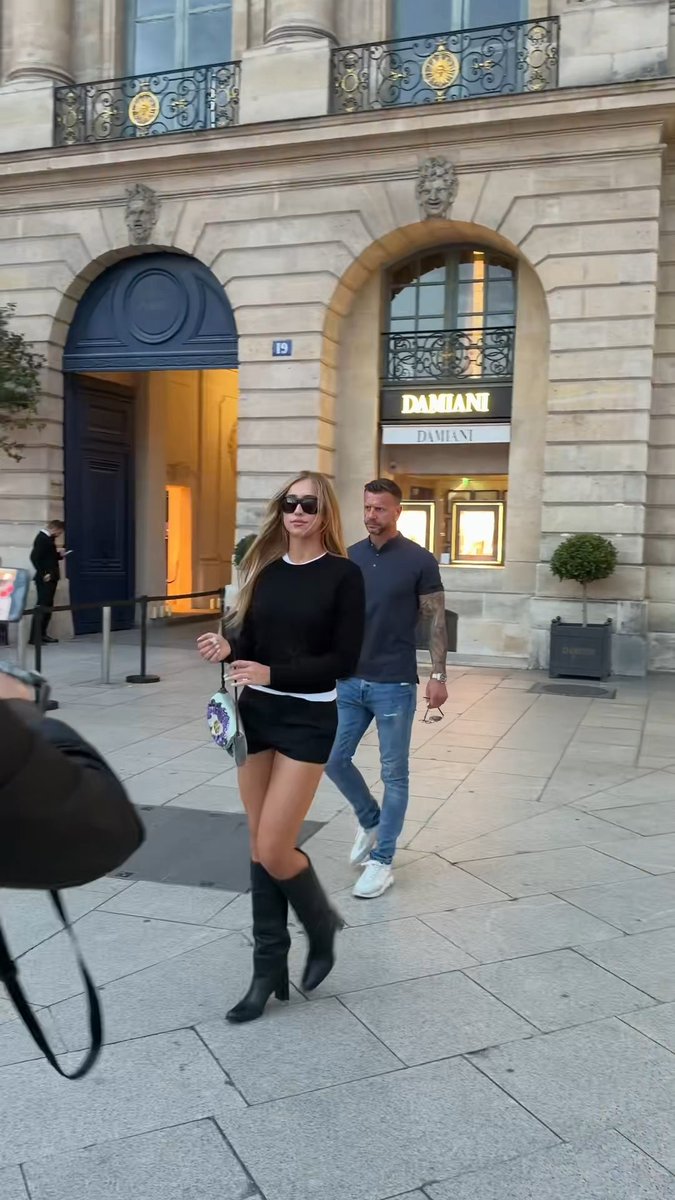 A blonde woman wearing sunglasses, a black sweater, black mini skirt, and black knee-high boots walking on a Parisian street past a Damiani jewelry store at address 19, with a man in a navy polo shirt and jeans following behind her.