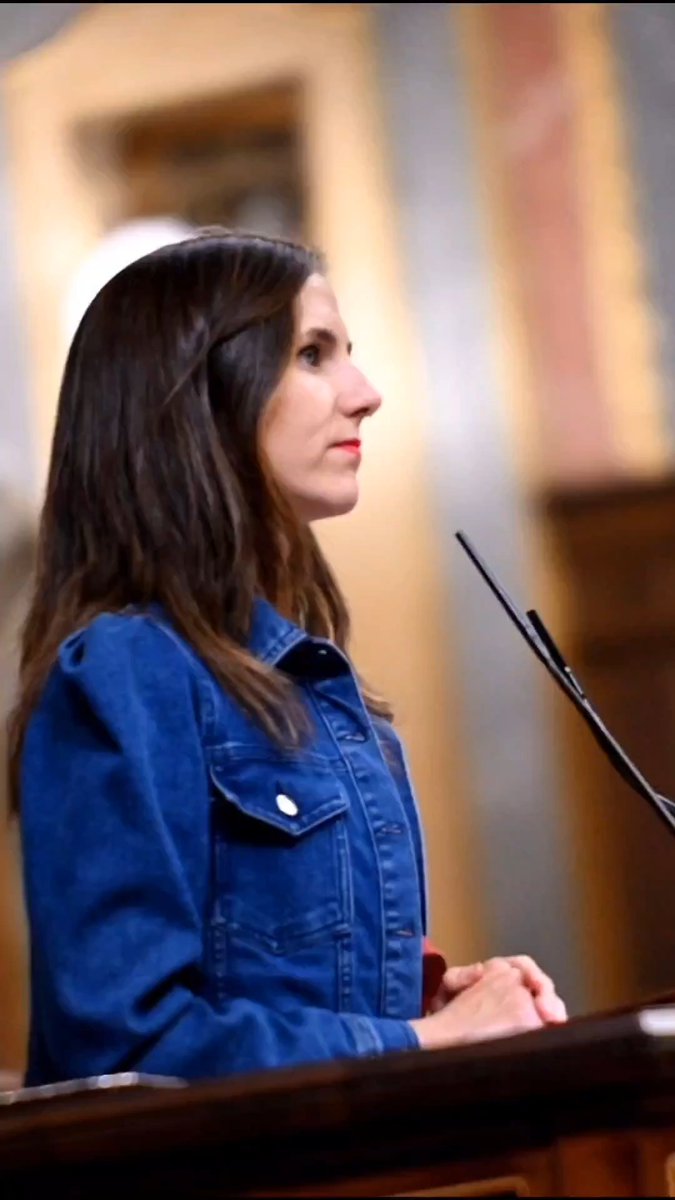 A side-profile shot of a woman with long brown hair wearing a blue denim jacket, standing at a podium with a microphone in what appears to be a government building.
