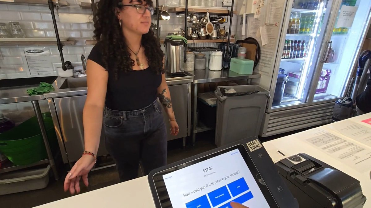 A woman stands behind the counter of what appears to be a small café, with a tip screen in the foreground displaying "$17.32" and the prompt "How would you like to receive your receipt?" along with a "NO TIP" option visible on the touchscreen. A small "HERE? TO GO?" sign sits on the counter next to the point-of-sale terminal, while behind her a refrigerated display case holds bottled drinks. The image captures the exact moment referenced in the post—the employee is distracted and looking away as the tip screen awaits selection.