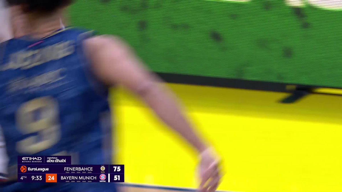 A blurry basketball player in a dark blue jersey with number 9 walks past a green electronic display screen during a EuroLeague game. The scoreboard shows Fenerbahce leading Bayern Munich 75-51 with 9:33 remaining in the 4th quarter, consistent with the recent fast break scoring play by Johnny Juzang for Fenerbahce.
