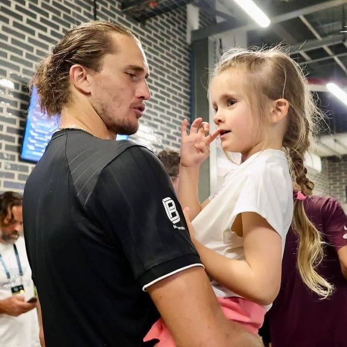 A tall man with long hair holds a young blonde girl with a braid who is touching her face with her finger; he wears a black shirt with 'uhlsport' and 'we united' text.