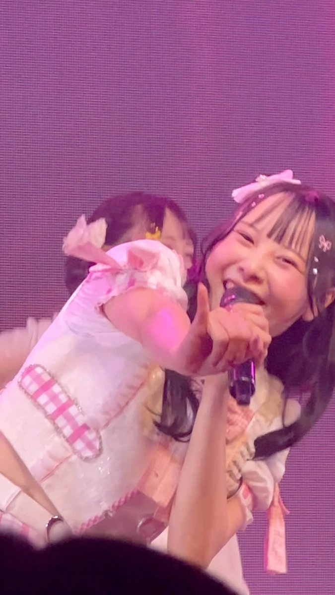 A young performer in a frilly pink and white outfit sings into a microphone while holding a doll on stage under purple lighting.
