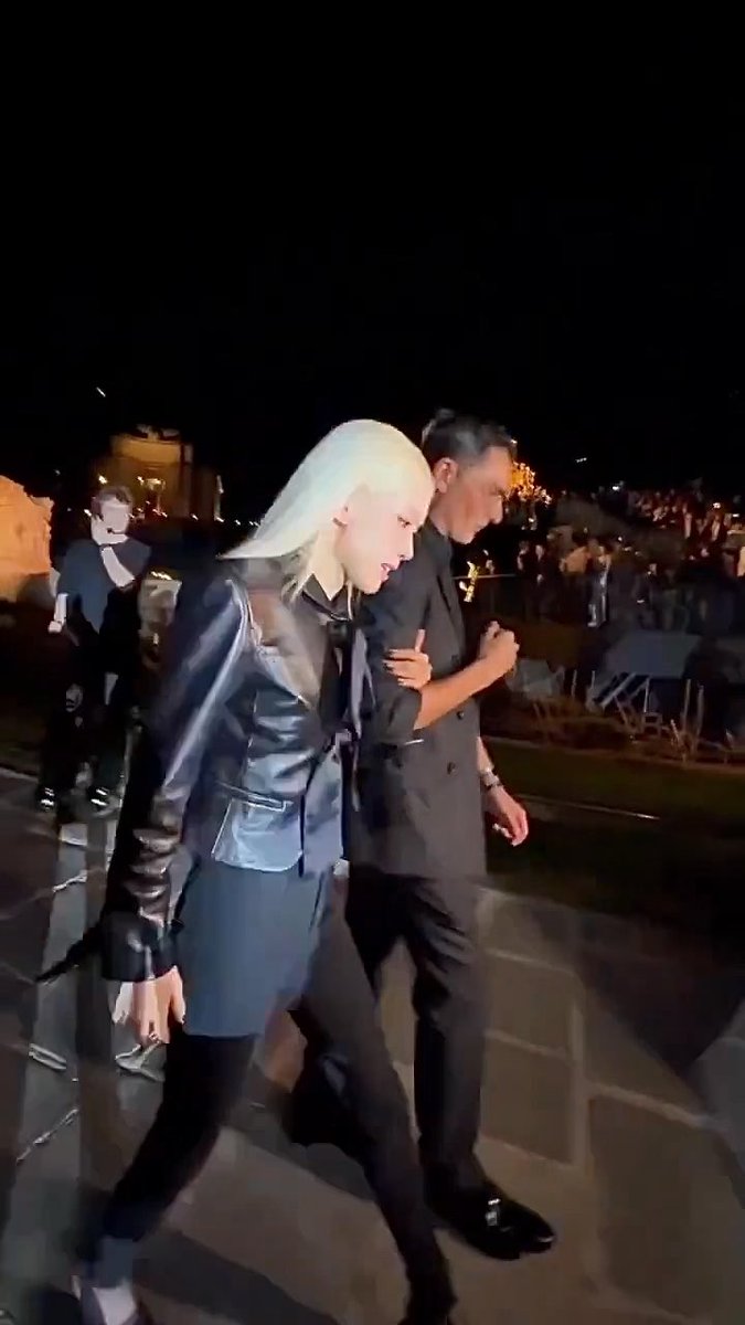 Nighttime candid photo of a blonde-haired person in a black leather jacket walking alongside a man in a dark suit, with a crowd seated in the background at an outdoor evening event.