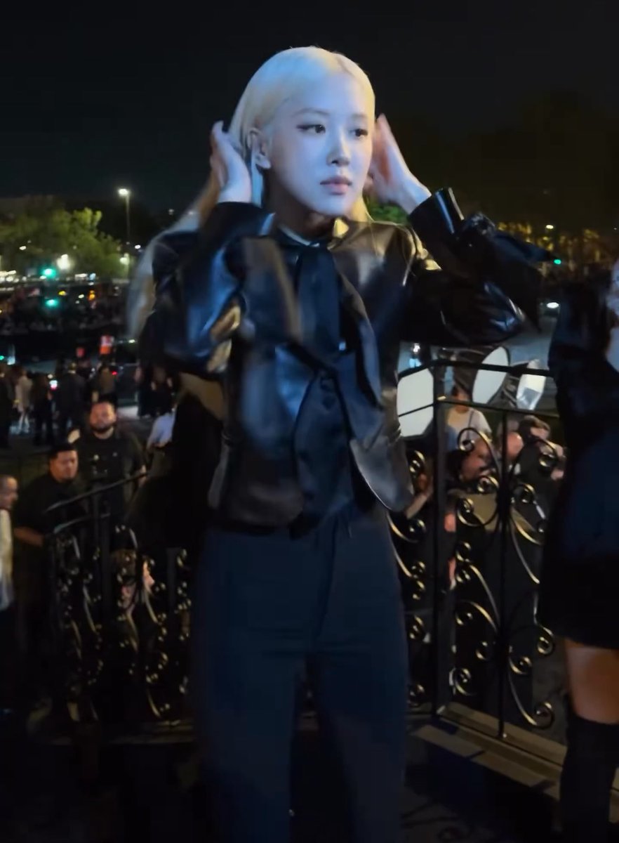 A blonde woman wearing a black leather jacket stands behind a decorative black metal railing at night, holding her hands to her head with an ornate illuminated gate in the background.