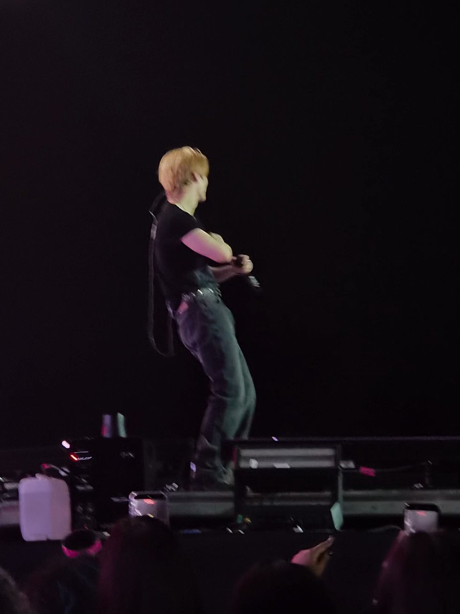 A performer with reddish-blonde hair, wearing a black t-shirt and dark jeans, stands on a dimly lit stage holding a microphone.