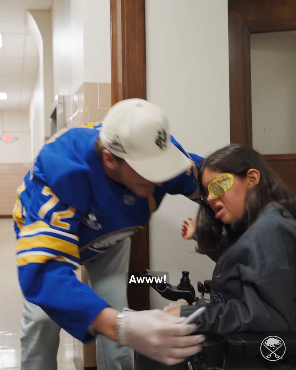A man in a Buffalo Sabres jersey (#72, Tage Thompson) leans down warmly while wearing gloves to interact with a student in a wheelchair at St. Mary's School for the Deaf, where the caption "Awww!" highlights the tender moment captured during the team's Community Day visit.