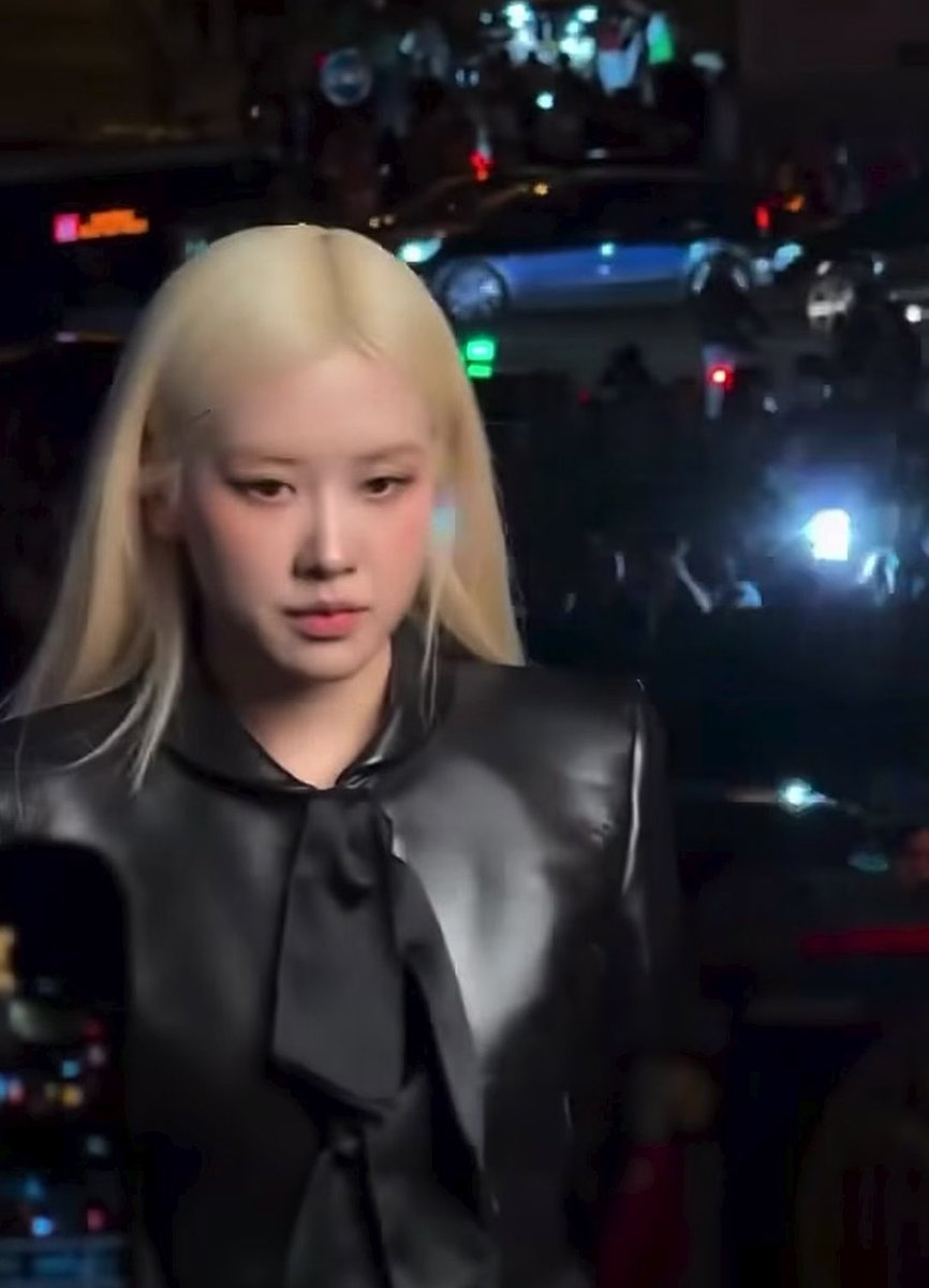 A blonde-haired woman wearing a black leather top with a bow tie is captured in what appears to be a candid moment at an event, with lighting visible in the background.