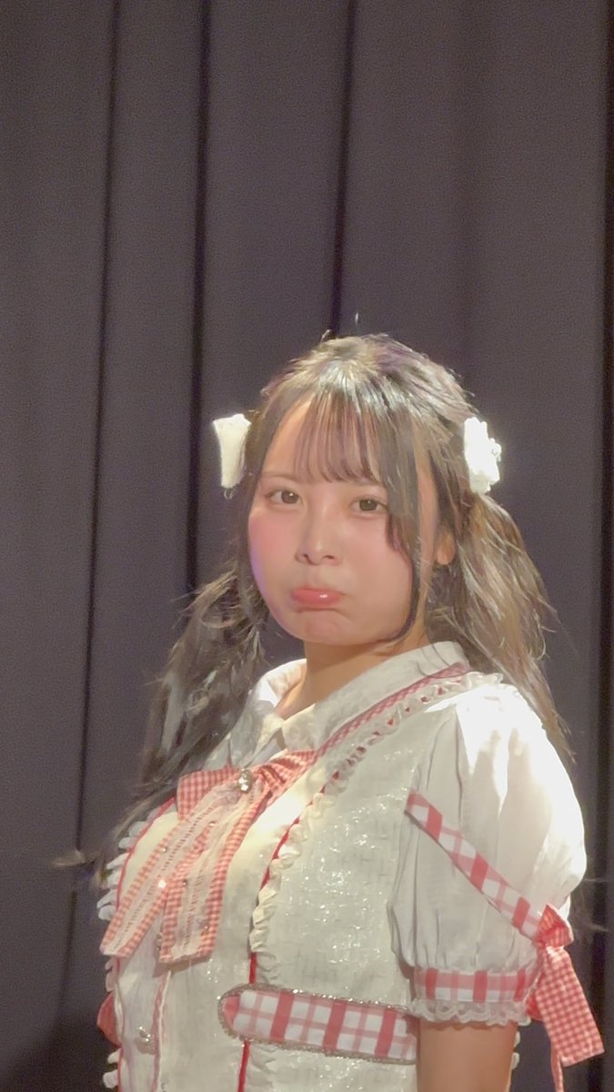 A young woman with long hair and white hair accessories wears a white and red gingham dress, looking at the camera against a black curtain backdrop.