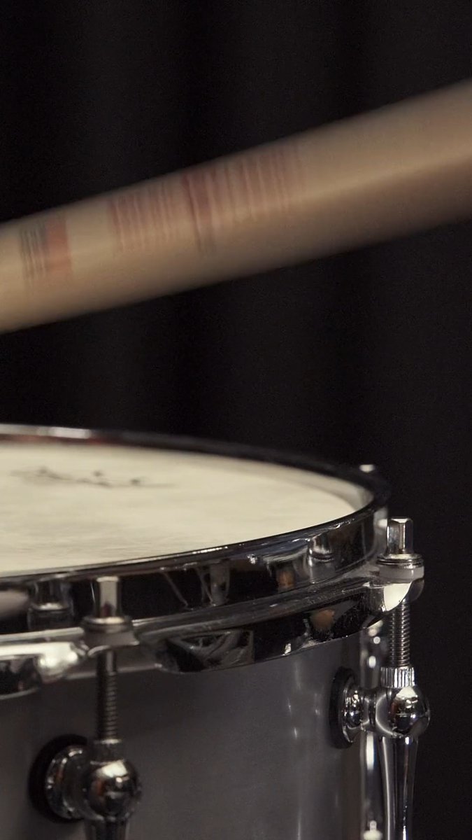 A close-up shot shows a drumstick in mid-air above a snare drum, capturing the moment just before a strike.